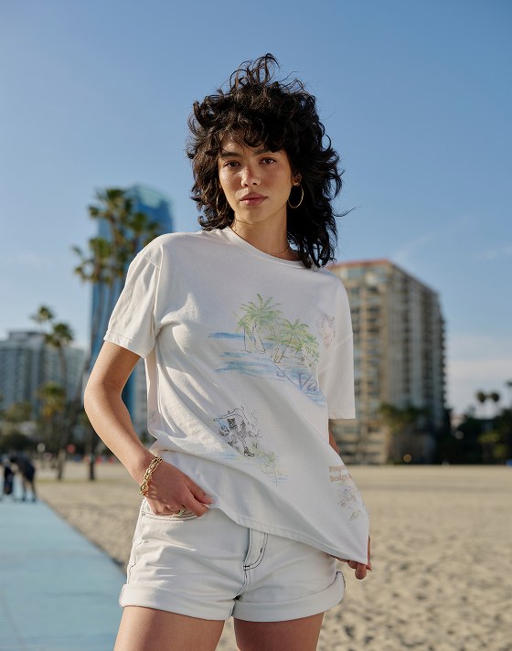 A young woman in a Vintage St. Vans shirt poses for the camera on a beach walkway with palm trees in the background.