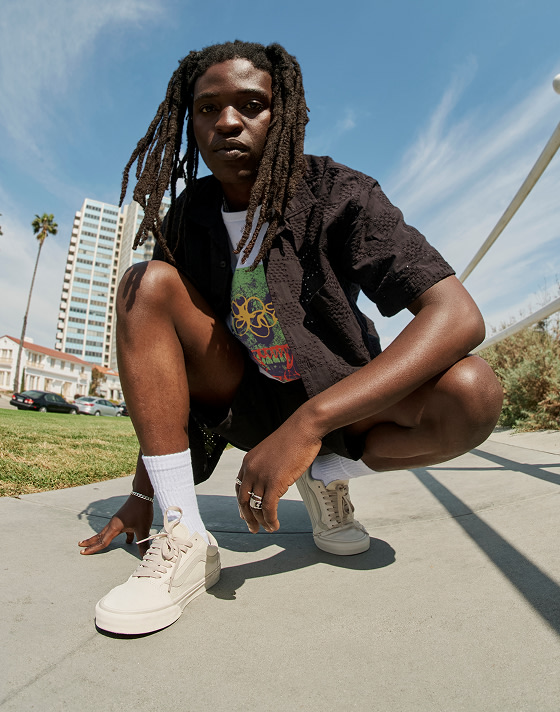 A young man squats on a sidewalk and looks into the camera wearing black Vans clothes and Warm Beige Old Skool.