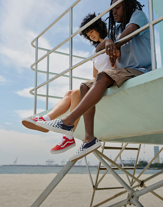 Two friends sit on a lifeguard tower kicking their red Old Skool and blue Checkerboard Classic Slip-On in the air with the beach behind them.