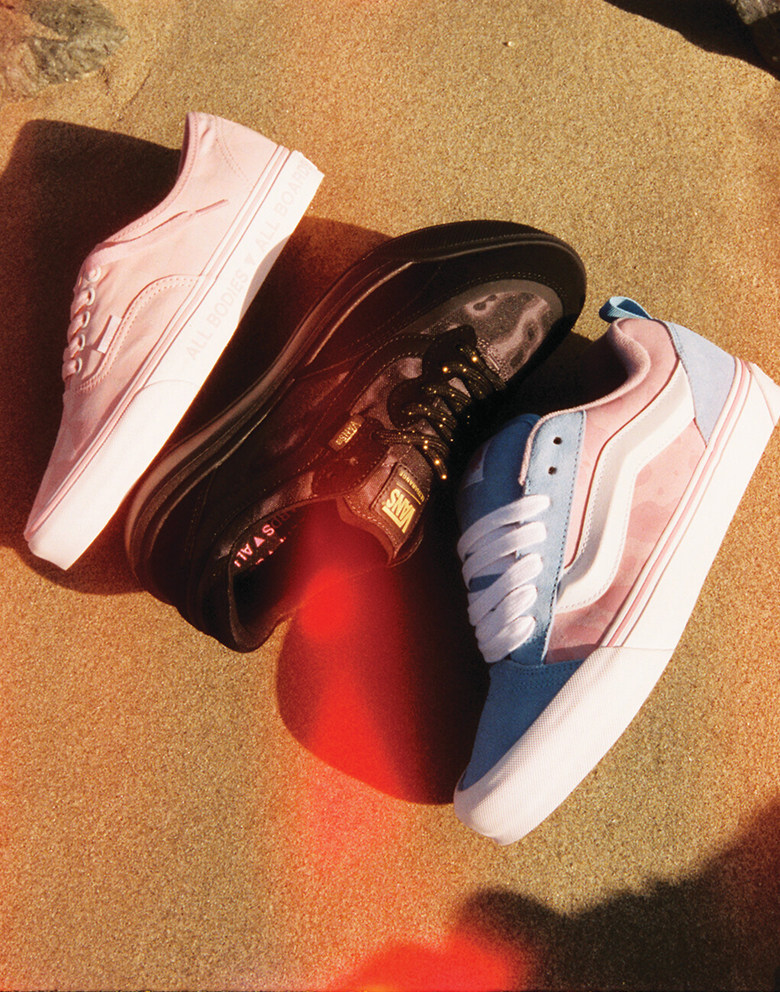 An Authentic shoe, an UltraRange 2.0 shoe, and a Knu Skool shoe from the Vans x Queer Surf collection lay on the sandy beach under a lens flare.