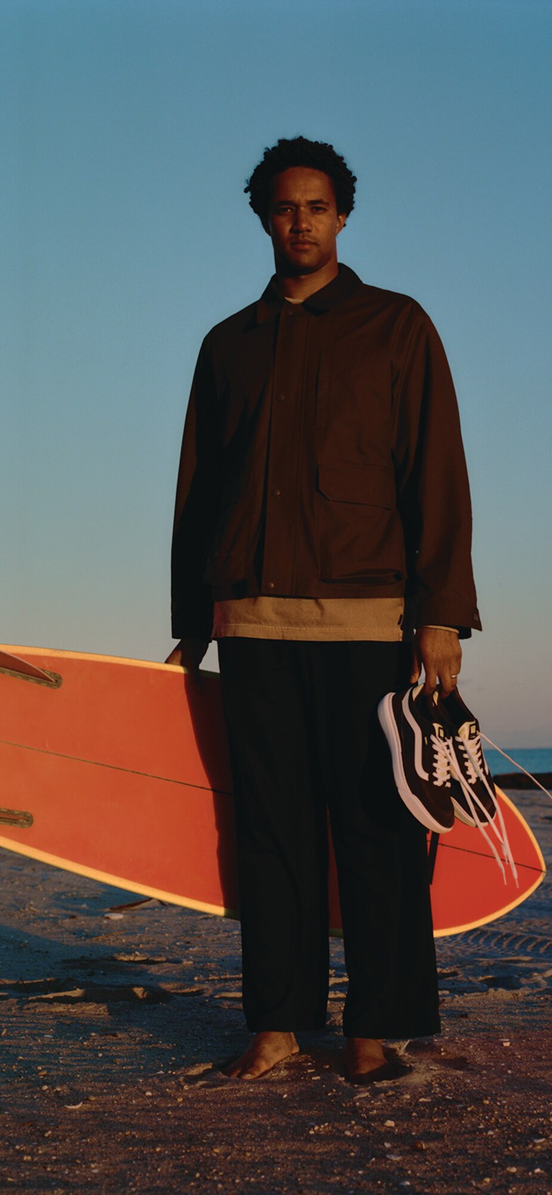 A man poses at the beach holding black and white UltraRange 2.0 shoes in one hand and a surfboard in the other during golden hour.