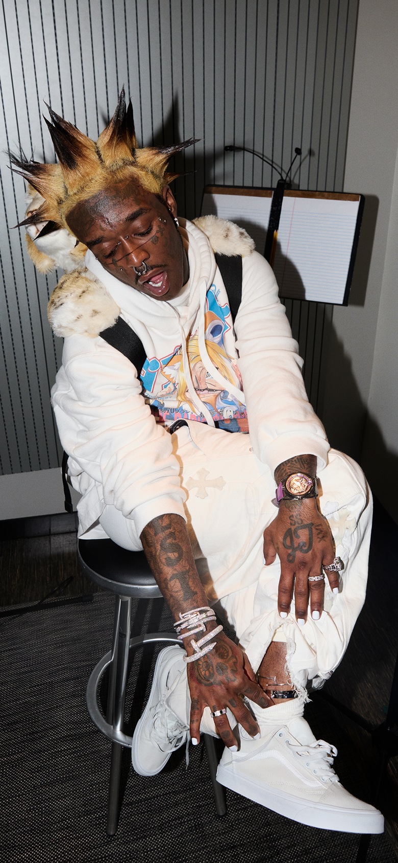 Lil Uzi Vert wears white pants and a white hoodie, showing a silver anklet over his white Old Skool shoes in his studio.