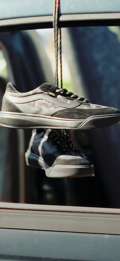 A black and gold yellow Vans X Queer Surf UltraRange 2.0 Shoe hangs from a car window near the beach.
