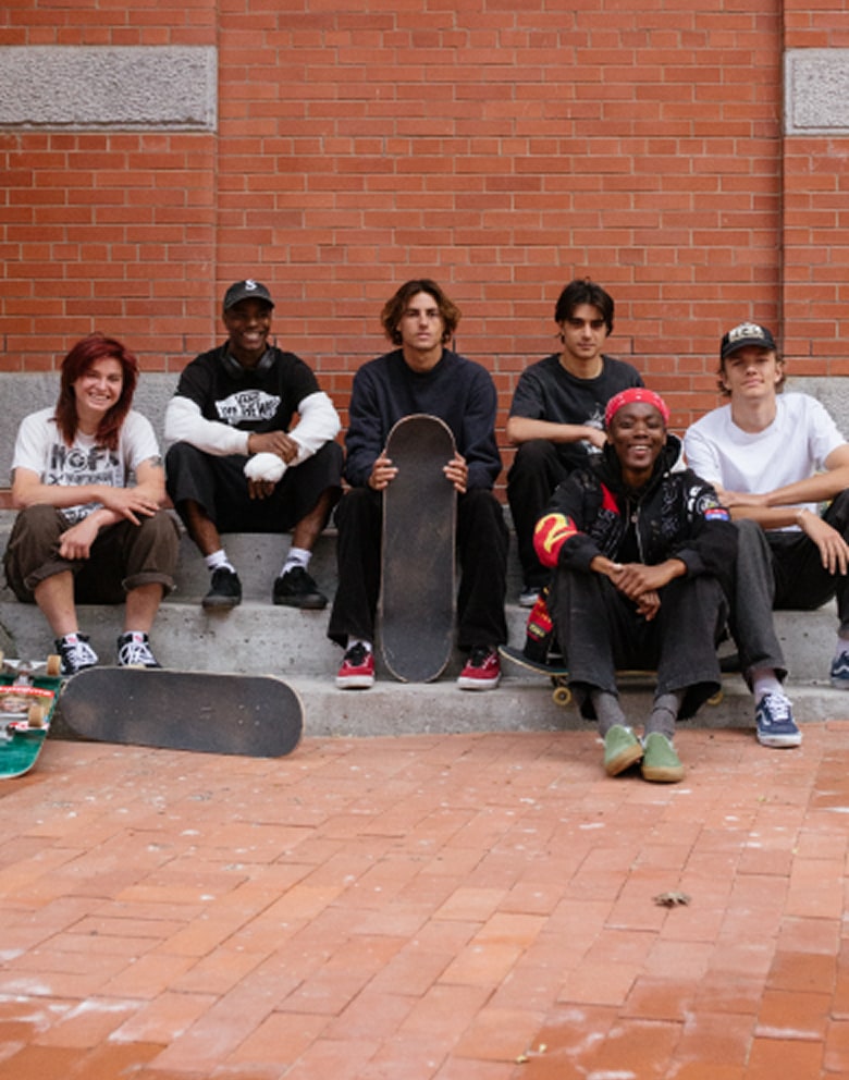 A group of Vans skaters sit and smile in front of a brick building with Curren Caples in the center holding a skateboard and wearing Curren shoes.