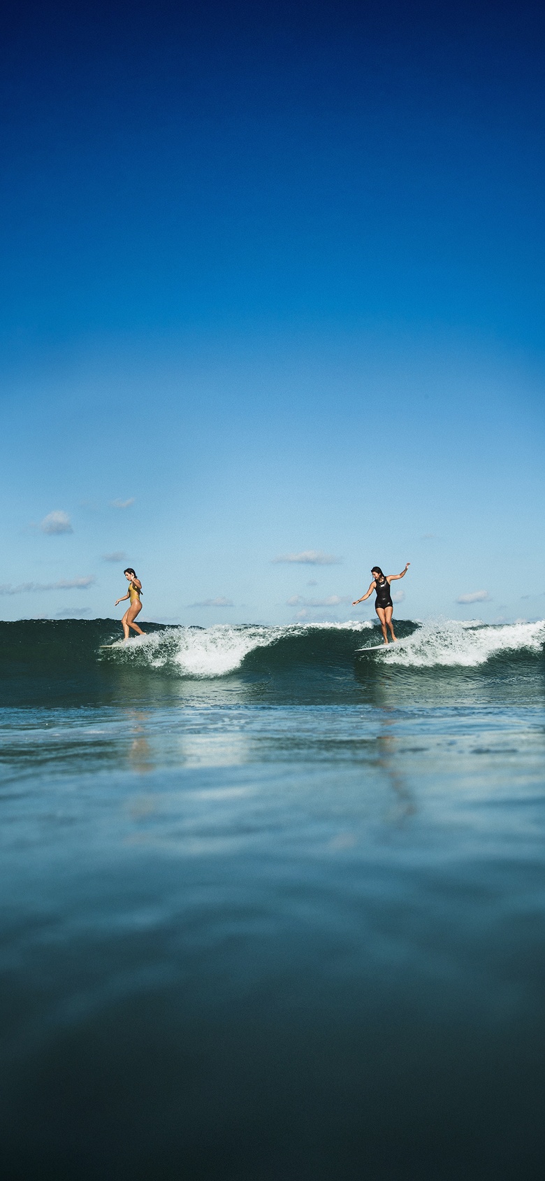 Vans surfers Lola Mignot and Karina Rozunko surf a wave together on a clear day.