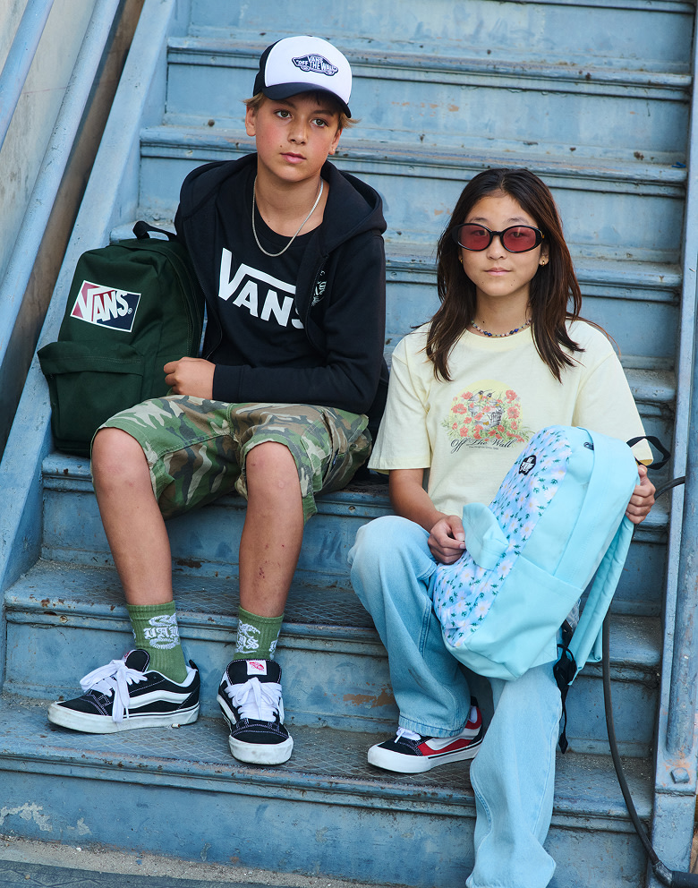 A boy and girl walk down an alley wearing Vans clothes and backpacks wearing Knu Skool shoes.