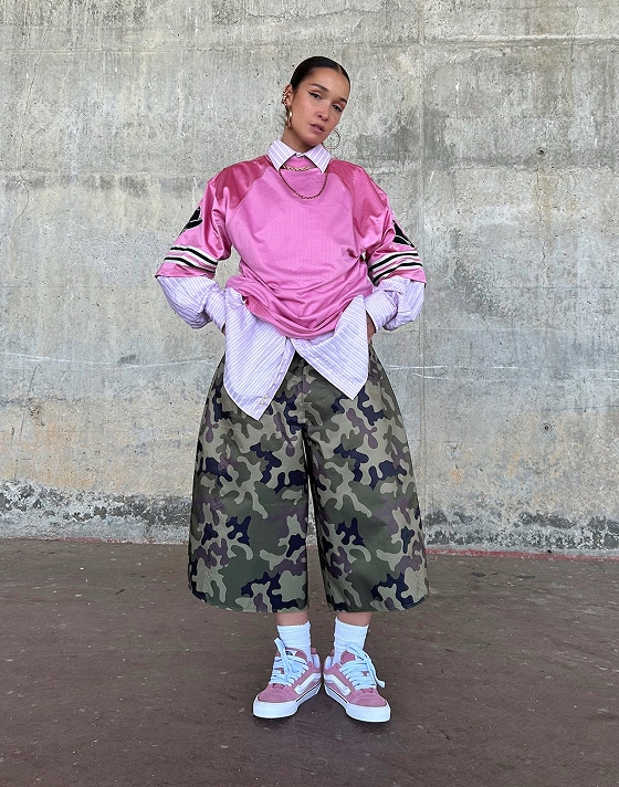 A young woman poses for the camera in a pink football jersey, baggy camo pants, and pink and white Knu Skool shoes.