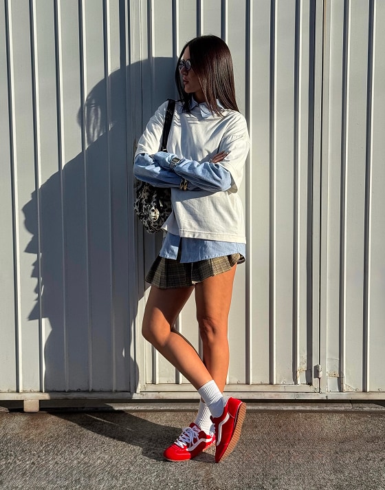 A young woman leans up against a metal fence with her arms crossed wearing a baggy shirt, a skirt, and Racing Red Super Lowpro.