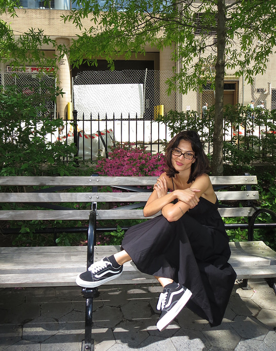 A young woman in a black dress smiles at the camera and sits on a park bench wearing black and white Old Skool shoes.