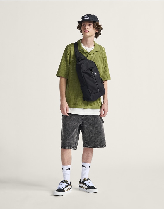 A young man poses in a photography studio wearing the Feldman Sweater Shirt, Drill Chore Loose Denim 22-inch Shorts, and Warp Ripstop Sling Bag.