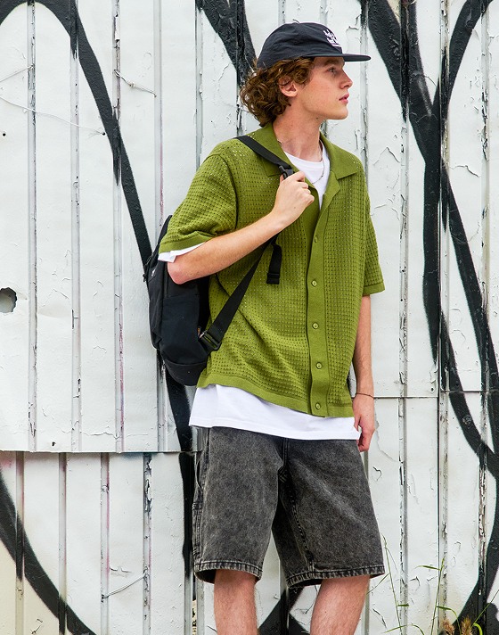 A young man leans up against a wall wearing the Feldman Sweater Shirt and Drill Chore Loose Denim 22-inch Shorts.