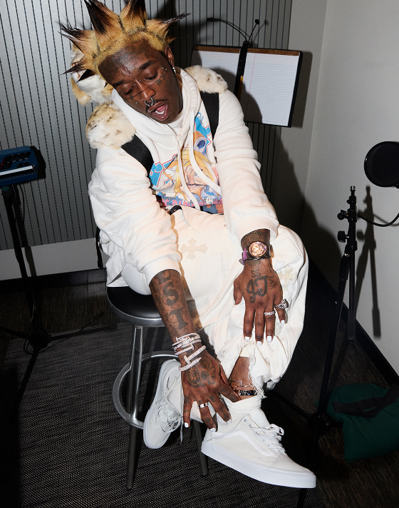 Lil Uzi Vert wears white pants and a white hoodie, showing a silver anklet over his white Old Skool shoes in his studio.