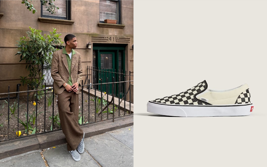A young man leans against a railing under a building in New York City in a baggy brown houndstooth suit while wearing Checkerboard Classic Slip-On.
