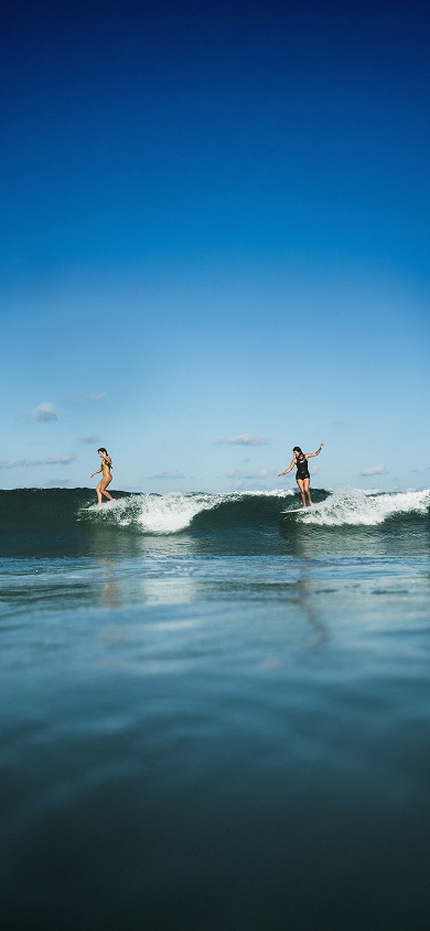 Vans surfers Lola Mignot and Karina Rozunko surf a wave together on a clear day.