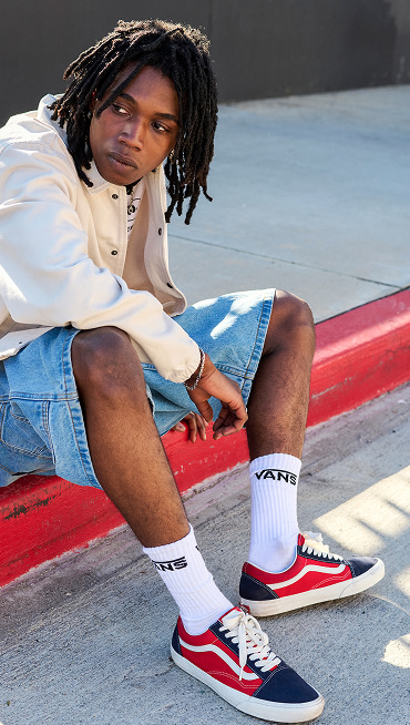 A person sits on a sidewalk curb relaxing, wearing the Vans Old Skool shoe in the Navy Blue/Red color with denim shorts and an ivory jacket.