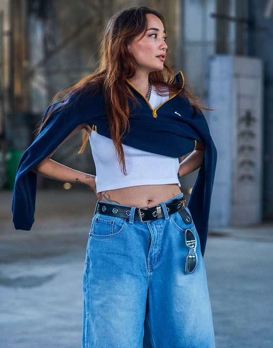 A woman poses in a warehouse wearing a Vans navy half-zip, cropped white rib tank, wide leg jeans, and a black leather belt.