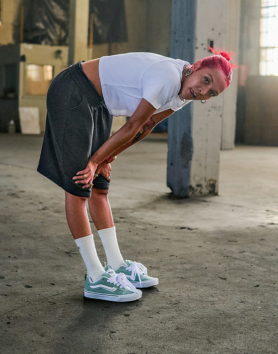 A person poses in a warehouse wearing baggy shorts, a cropped white tee and Vans Knu Skool shoes in grey mint green.