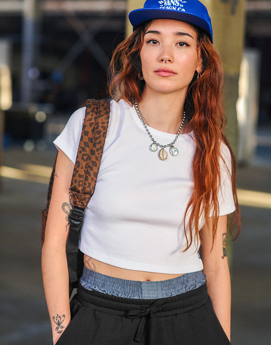 Waist up portrait of a woman wearing a cropped white tee, true blue Vans HOV Snapback Hat and the Old Skool Classic Backpack in Dachshund Brown.