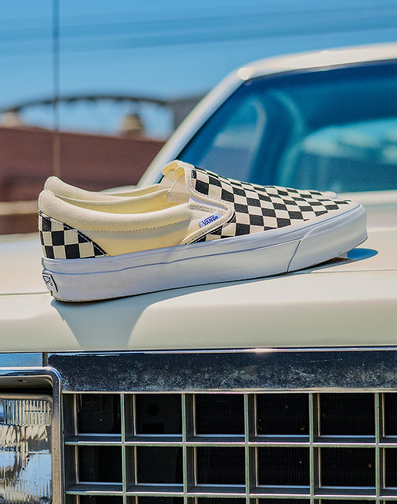 The Vans Classic Slip-On shoe in the iconic Checkerboard Black/Off White color rests on the hood of a vintage car in the bright sun.
