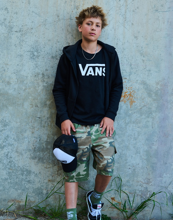 A boy poses against a wall wearing a black Vans hoodie, camo shorts, Knu Skool shoes and holds the Classic Patch Curved Bill Trucker Hat.