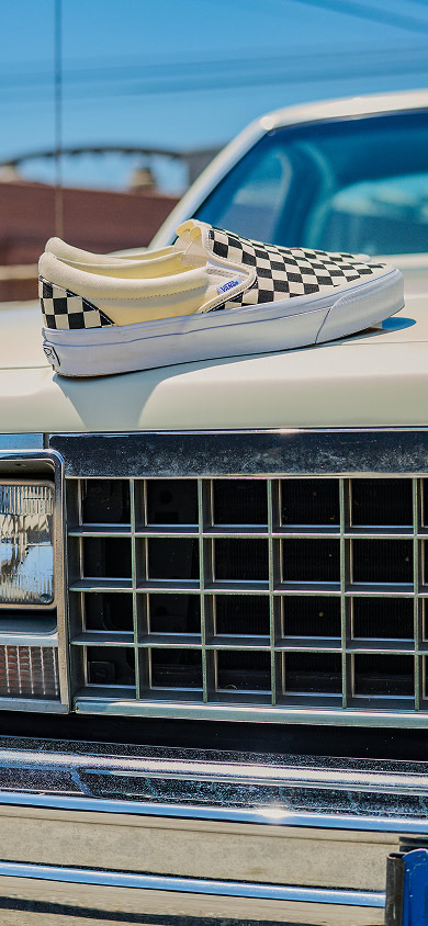Black and white checkerboard slip-on Vans sitting on the hood of a car outdoors.