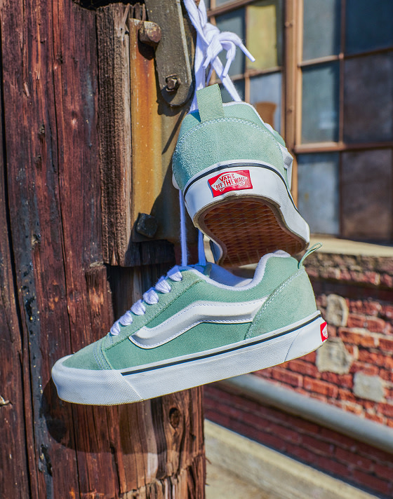 Close up of grey mint green Knu Skool shoes hanging from a weathered wood post, with a brick wall in the background.