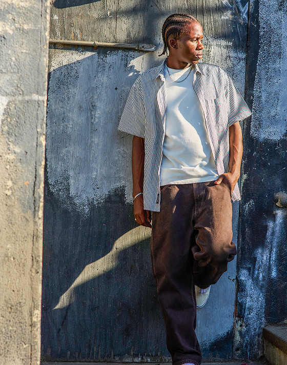 A man poses during golden hour against a half-painted wall wearing the Lawson Stripe Button Up Shirt over an ivory Vans tee shirt, and brown pants.