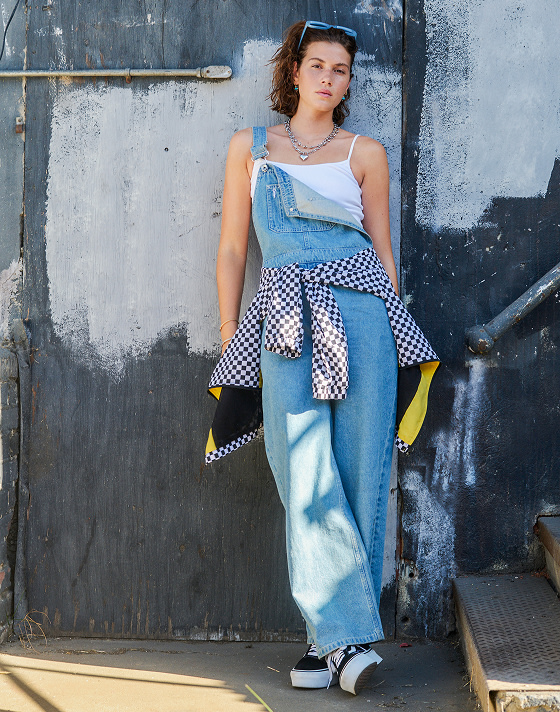 A woman leans against a half-painted wall outside, wearing denim overalls, a checkered windbreaker and classic Vans Old Skool shoes.