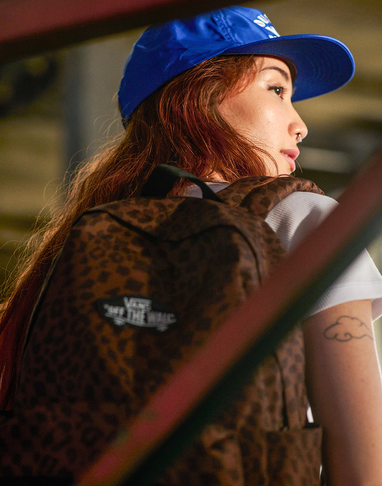 Waist up portrait of a woman wearing a cropped white tee, true blue Vans HOV Snapback Hat and the Old Skool Classic Backpack in Dachshund Brown.