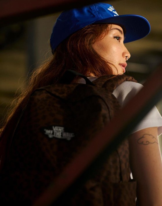 A woman looks over her shoulder in a warehouse wearing Vans clothes and a Dachsund Brown Old Skool Classic Backpack.