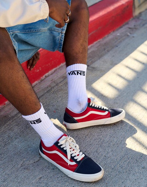A waist-down shot of a man sitting on a curb wearing red and blue Old Skool, denim shorts, and white Vans crew socks.