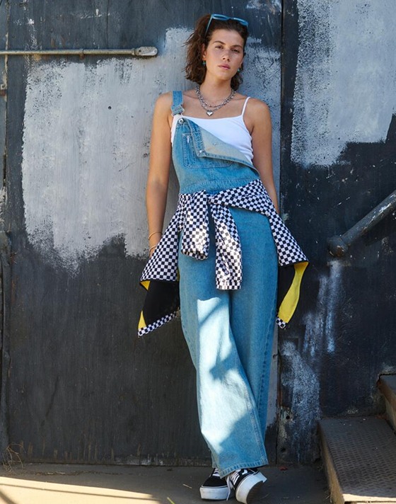 A woman leans up against a wall wearing denim blue Sirelle Overalls, a white tanktop, and black and white Platform Old Skool.
