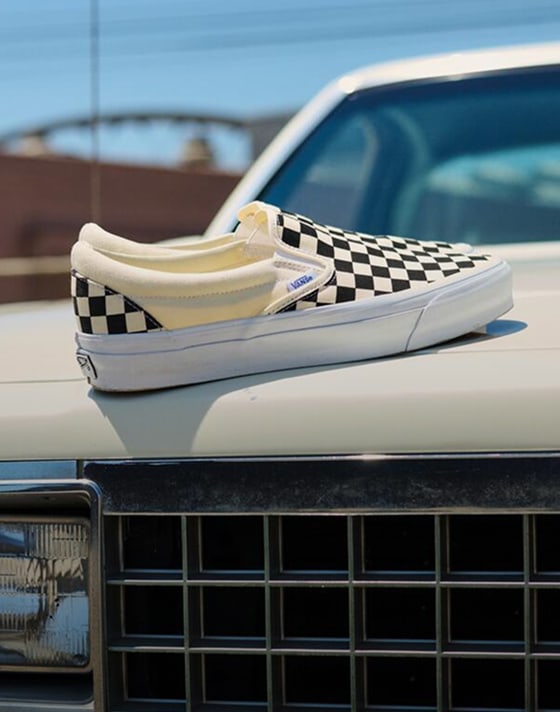A pair of black and white Checkerboard Classic Slip-On rest on the hood of a white car in the afternoon sun.