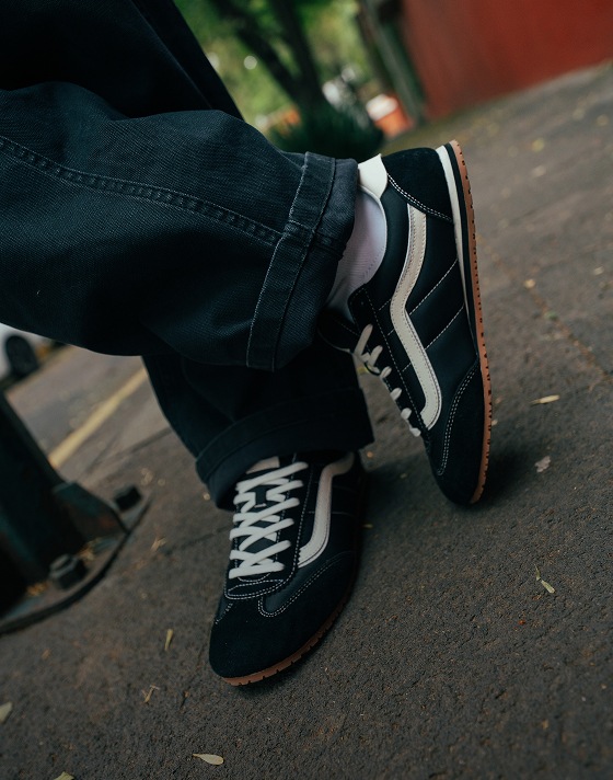 A close-up of someone leaning up against a pole wearing black and white Super Lowpro shoes and dark denim jeans.