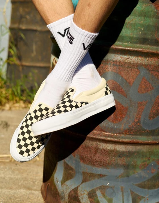 A person crosses their legs while sitting on an oil drum wearing white Vans crew socks and black and white Checkerboard Classic Slip-On.