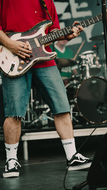 A guitarist plays on stage wearing denim shorts, white Vans crew socks, and black and white Old Skool.