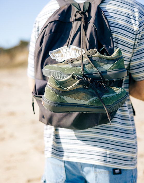 A pair of coal brown UltraRange 2.0 hang by their laces from a Old Skool Classic Backpack being worn on the beach.