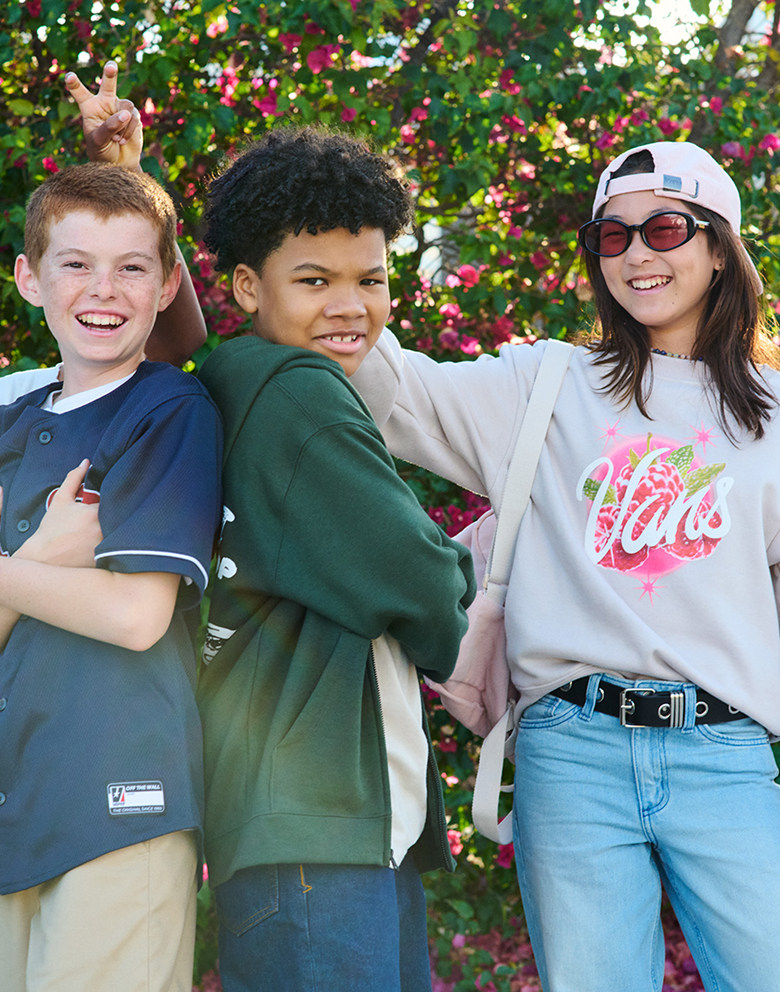 A group of young kids smile and pose for the camera in Vans clothes and backpacks in front of a rose bush.