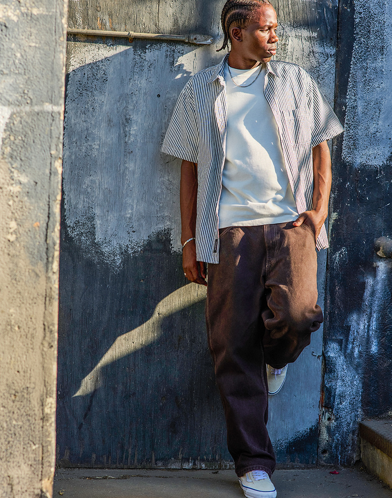 A man leans on a wall wearing a blue and white Lawson Stripe Button Up Shirt and brown jeans.