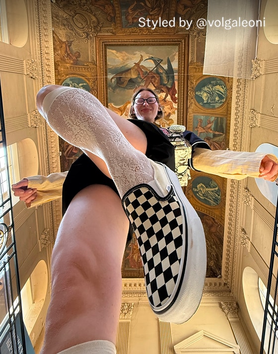 A woman steps over the camera wearing knee-high socks and black and white Checkerboard Classic Slip-On inside a museum.