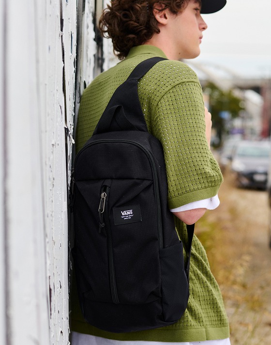 A boy leans up against a wall wearing a black Warp Ripstop Sling Bag and a Loden Green Feldman Sweater Shirt.