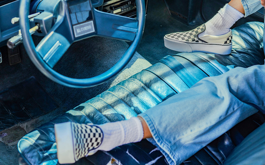 A waist-down shot of someone in a retro car wearing blue jeans and black and white Checkerboard Classic Slip-On.