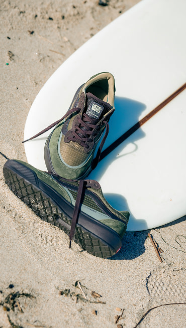 A pair of coal brown UltraRange 2.0 rests on a surfboard on a beach on a sunny day.