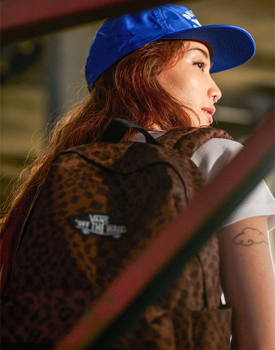 A woman looks over her shoulder in a warehouse wearing Vans clothes and a Dachsund Brown Old Skool Classic Backpack.