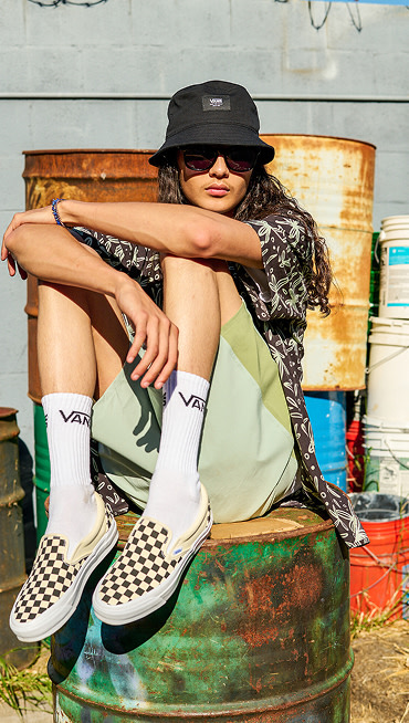 Close up of feet crossed by a person sitting on a graffitied oil drum, wearing white socks and checkerboard Classic Slip-On shoes.