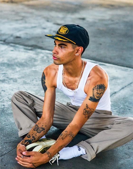 A man stretches on the concrete wearing desert taupe brown Authentic Chino Relaxed Pants and a black Scrambled Vans Snapback Hat.