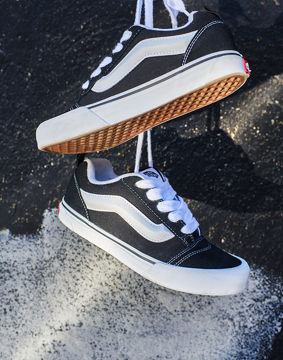 A pair of black and white Knu Skool hang by their laces against a graffiti-tagged concrete wall.