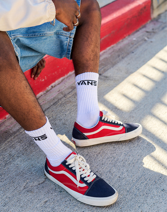 A young man looks down the street and sits on a curb in red and navy Old Skool paired with blue denim jeans.