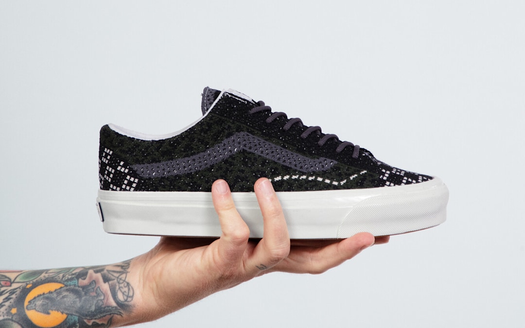 A Cheetah Dried Kelp Green Premium Old Skool is held up by someone with a tattooed arm in a photography studio.