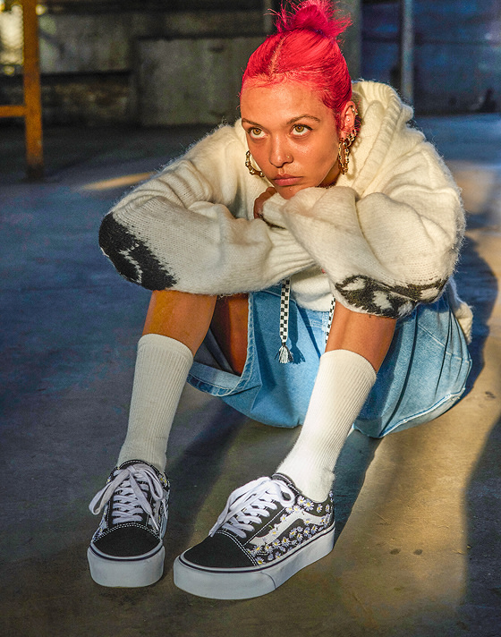 A woman with vibrant dyed red hair sits on the floor of a warehouse wearing denim shorts, white crew socks, and Old Skool shoes in the sunlight.